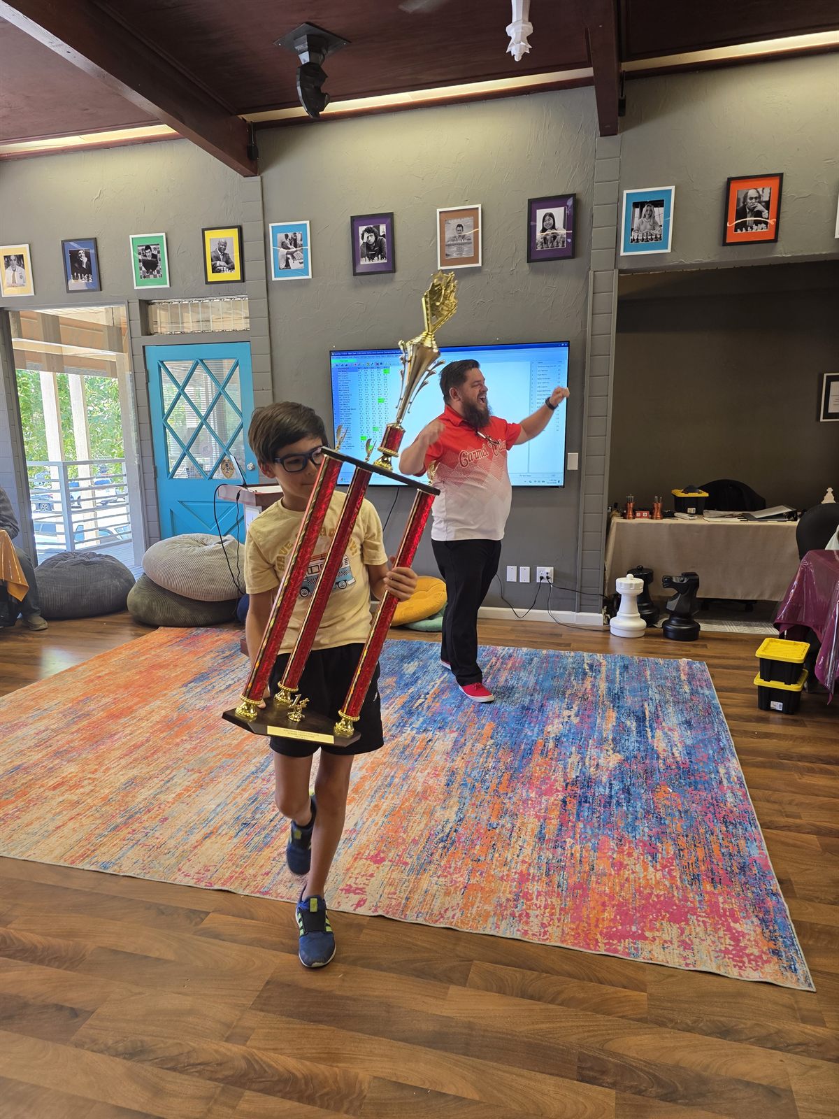 A young player carries a tall championship trophy across the room while Coach JB cheers in the background at Carmel Valley Chess Club