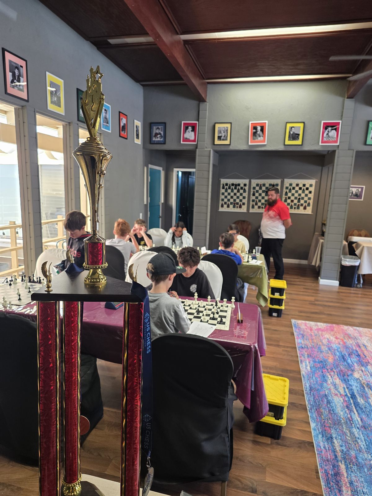 The tournament room at Carmel Valley Chess Club during a youth event, a championship trophy in the foreground and kids playing at the boards